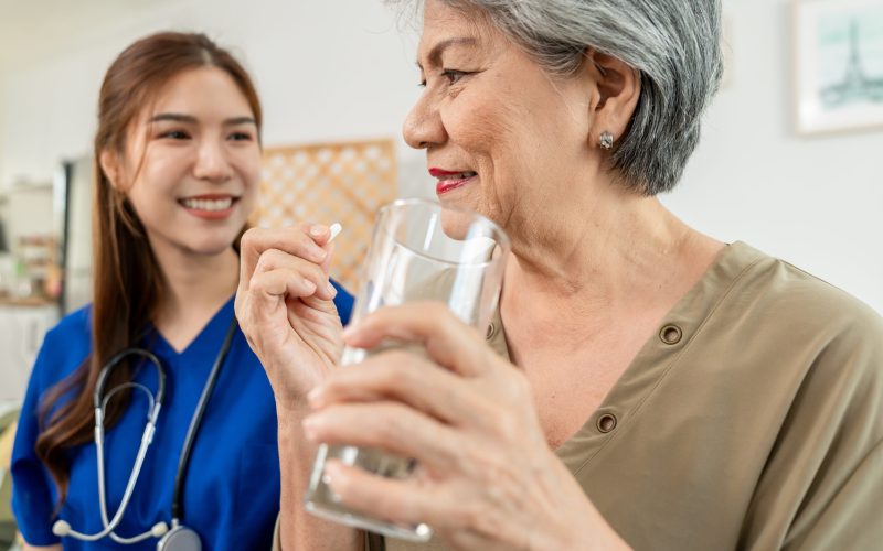 Asian caregiver give consultant to senior woman patient at nursing home. Attractive nurse carer giving medicine pills bottle, explain prescription to elderly mature grandmother in living room in house