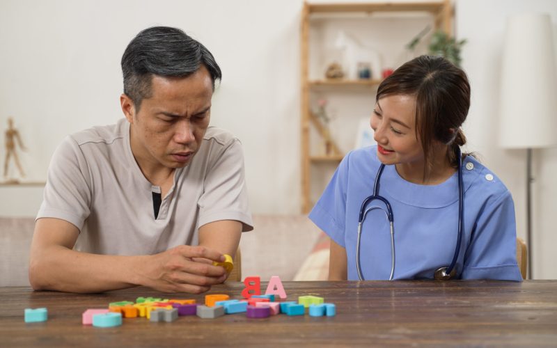 smiling asian female nursing aide giving instructions while helping the senior man go through physical treatment for dementia with color letter bricks at home